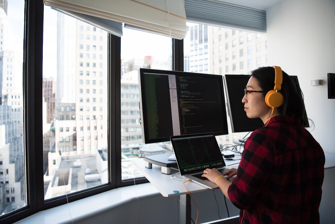 Person coding at a standing desk with laptop and dual monitors showing code, wearing yellow headphones, city skyline outside the window.