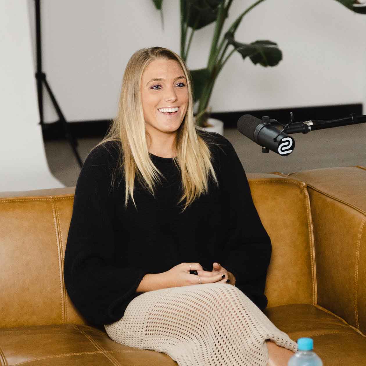Young woman smiling on a leather couch during a podcast interview, speaking toward a boom microphone; plant in background.