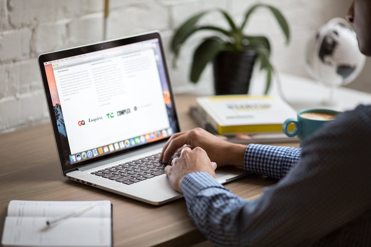 Hands typing on a laptop keyboard at a tidy desk with a notebook, potted plant, and coffee mug.