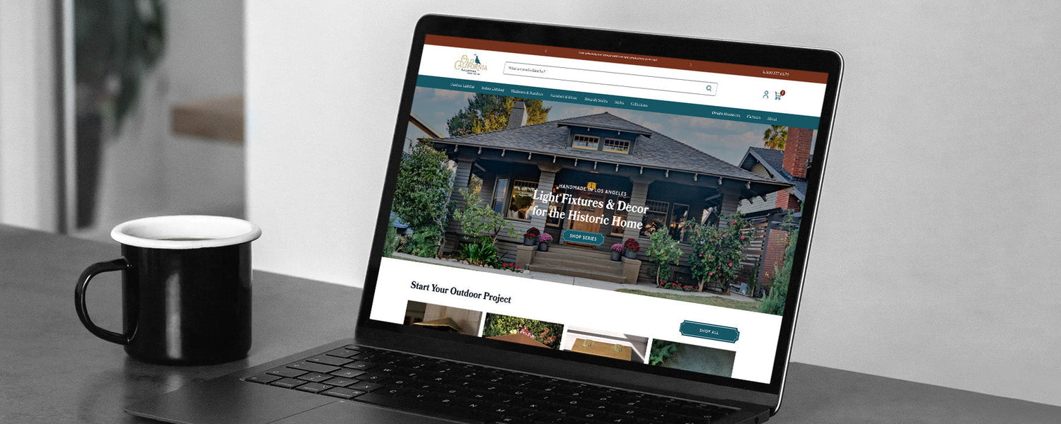 Laptop showing Old California homepage banner 'Light Fixtures & Décor for the Historic Home' with coffee mug beside it