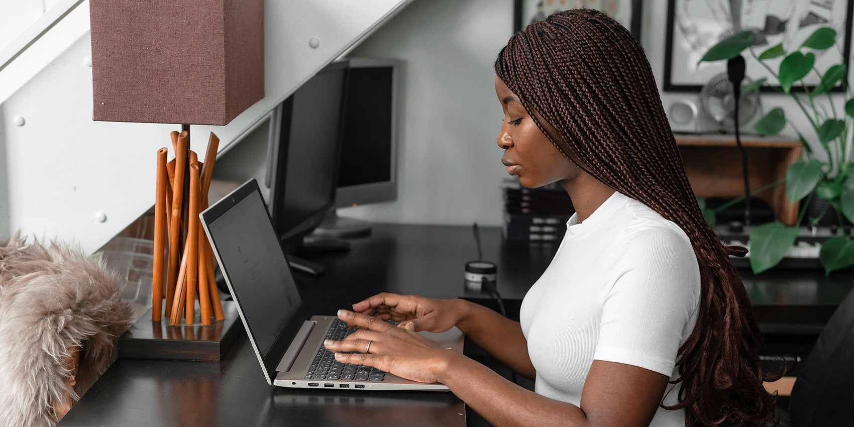 Young woman with long braids typing on a laptop at a desk, side view with lamp and potted plant in background.